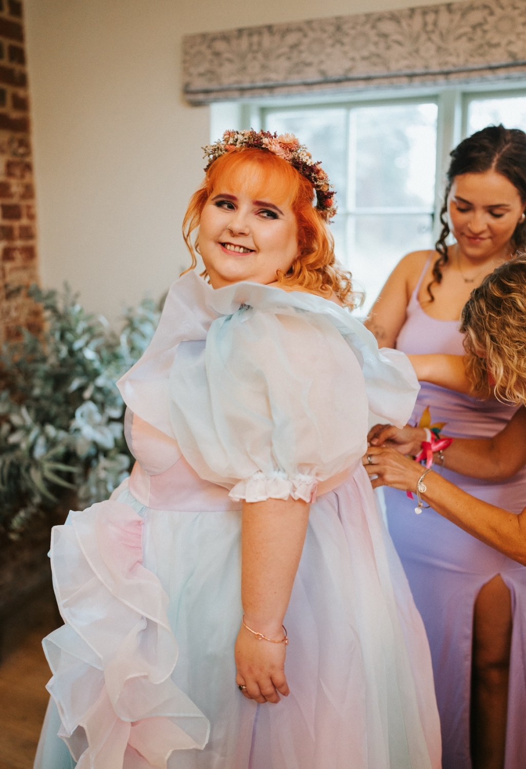 Lauren, a plus sized ginger, is standing, wearing a flower crown and a flowing pastel dress. Their family are fussing over the details in the background.